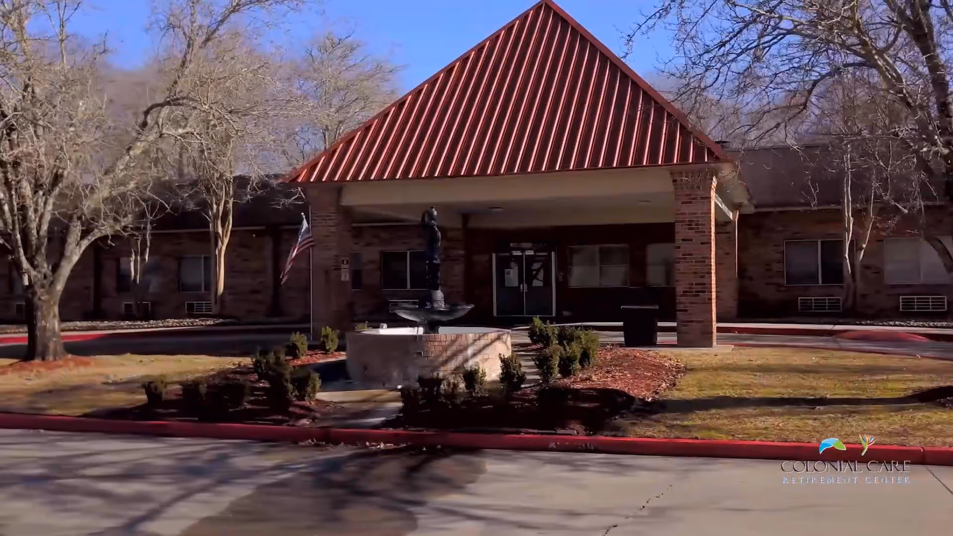 Exterior view of a retirement center building with a red metal roof over the entrance, brick walls, a circular driveway, and a small fountain surrounded by shrubs in front. Leafless trees are visible around the building under a clear blue sky.