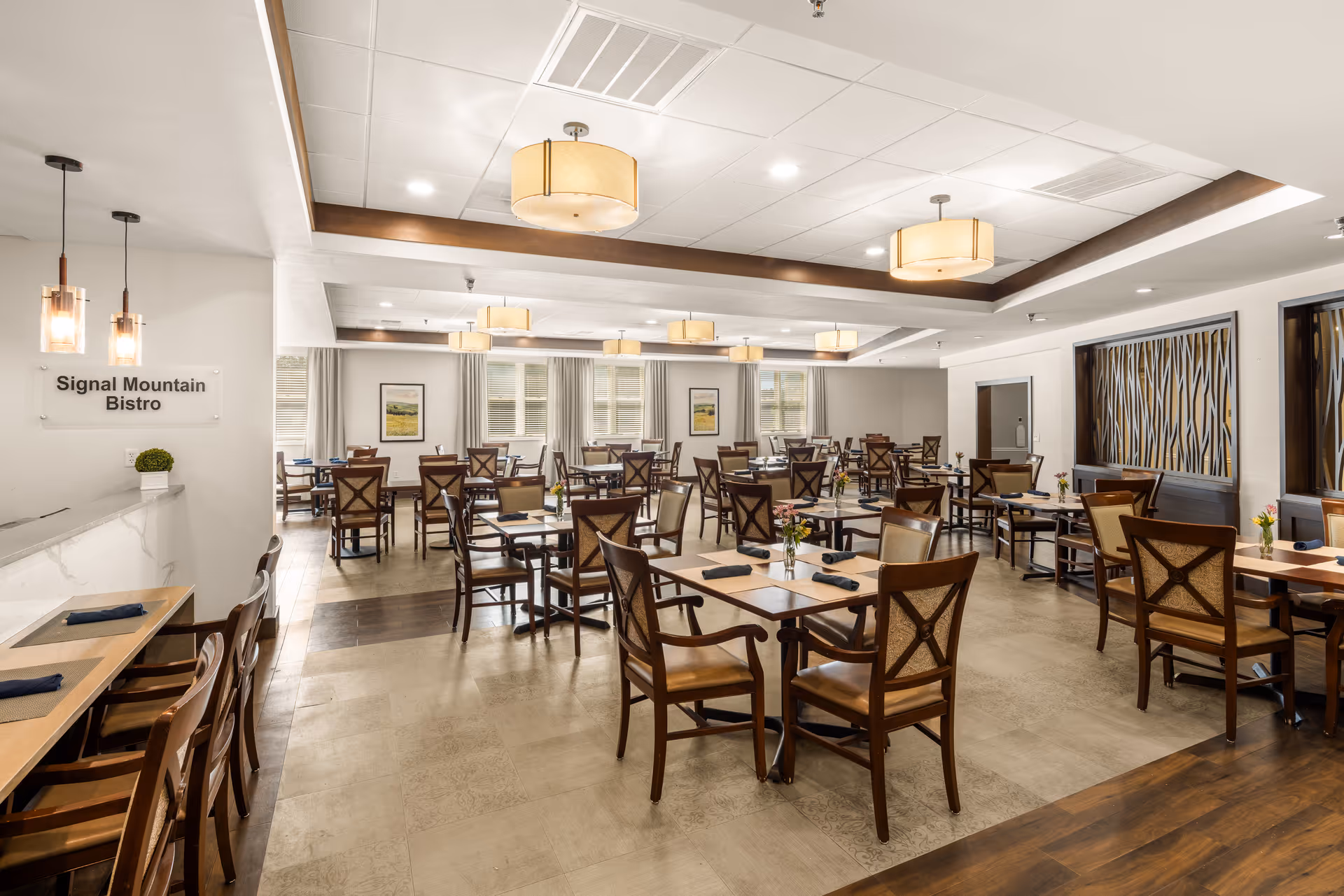 A spacious and well-lit dining room with multiple wooden tables and chairs arranged neatly. Each table has a small flower vase and black napkins. The room features modern ceiling lights, large windows with curtains, and decorative wall art. A sign on the left wall reads 'Signal Mountain Bistro'.