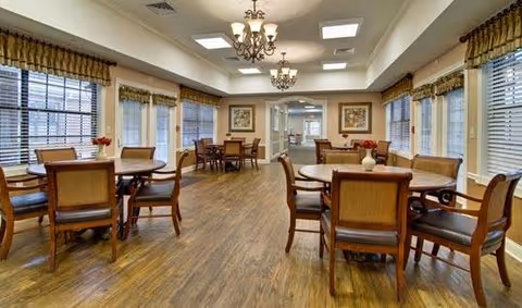 A spacious dining room with multiple round and rectangular wooden tables surrounded by chairs. The room has large windows with blinds and valances, chandeliers hanging from the ceiling, and framed artwork on the walls. The floor is wooden, and the room is well-lit with natural and artificial light.