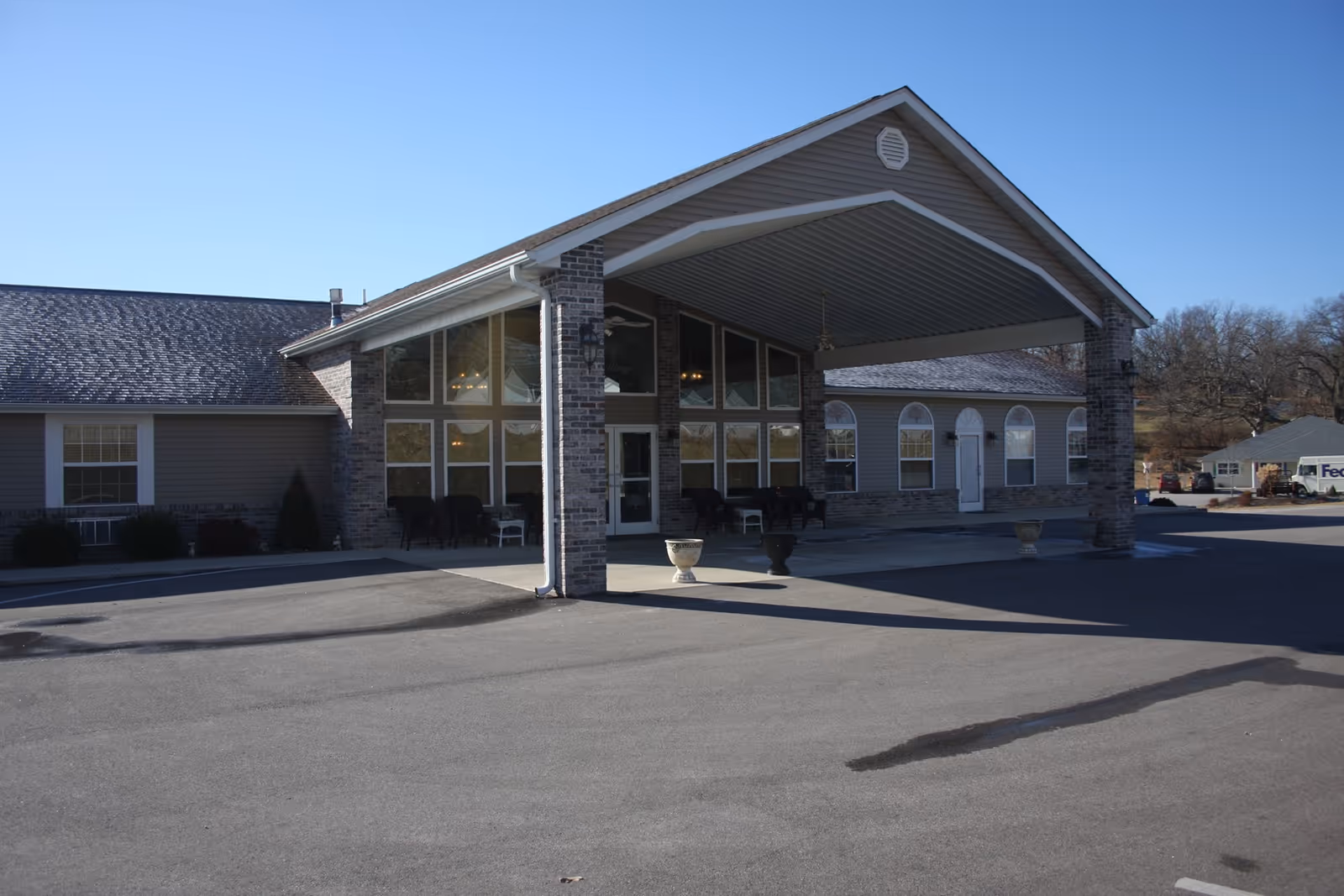 Exterior view of Crabapple Village Senior Estates showing the main entrance with a covered driveway supported by brick pillars. The building has large windows and a gray roof under a clear blue sky.