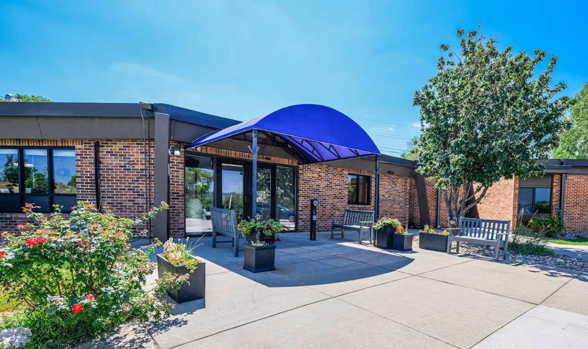 Outdoor entrance area of a brick building with a blue canopy over the doorway, surrounded by benches, potted plants, and trees under a clear blue sky.