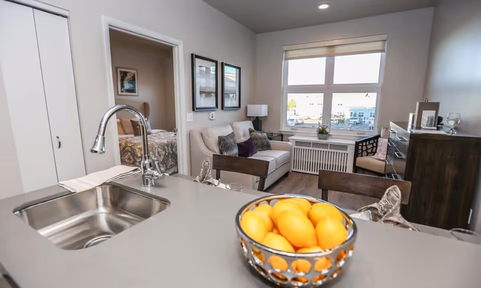 View of a modern apartment interior showing a kitchen counter with a stainless steel sink and a bowl of lemons in the foreground. Beyond the counter is a living area with a white sofa, decorative pillows, a window with a view outside, and a wooden dresser. A bedroom with a bed and artwork on the wall is visible through an open doorway.
