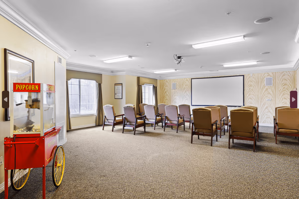 A small theater room with rows of cushioned chairs facing a large blank projection screen on the wall. There are windows with curtains on the left side and a red popcorn machine on wheels positioned near the entrance. The room has beige walls with a subtle pattern and carpeted flooring.