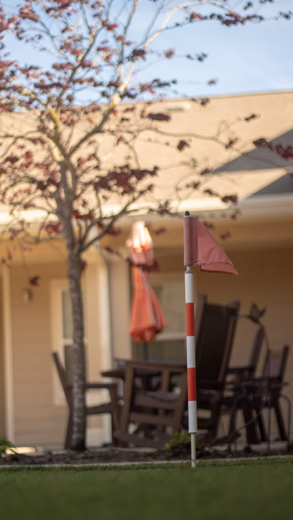 Red-and-white yard flag marker in a grassy outdoor courtyard with patio chairs, a red umbrella, and a tree in front of a building.