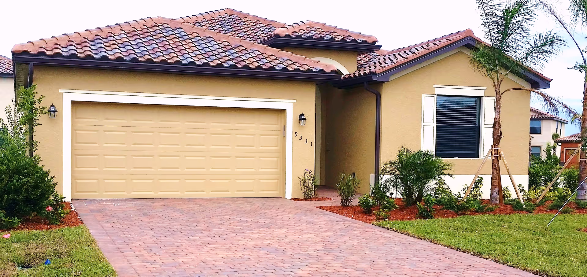 Front exterior of a single-story house with a tiled roof, two-car garage, paved driveway and landscaped yard.