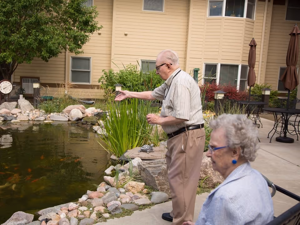 An elderly man and woman are outside near a pond with rocks and plants. The man is standing and reaching out towards the pond, while the woman is seated nearby. Behind them is a building with windows and outdoor tables with closed umbrellas.