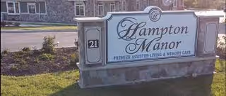 Outdoor view of a stone sign for Hampton Manor, a premier assisted living and memory care facility, located on a grassy area near a road with buildings in the background.