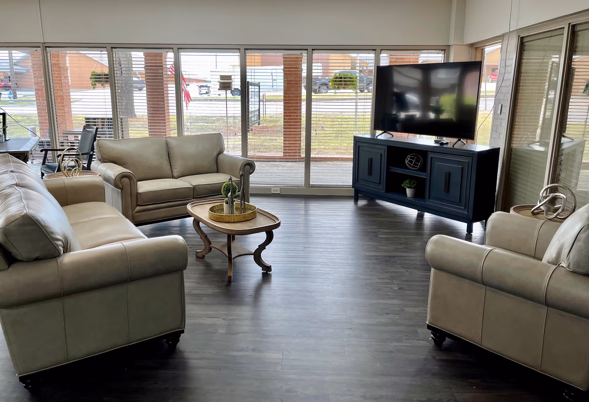 A bright living room with large floor-to-ceiling windows covered by horizontal blinds. The room features three beige leather sofas arranged around a wooden coffee table with decorative items. A flat-screen TV sits on a dark blue TV stand against the wall. The floor is dark wood, and outside the windows, a patio and some buildings are visible.