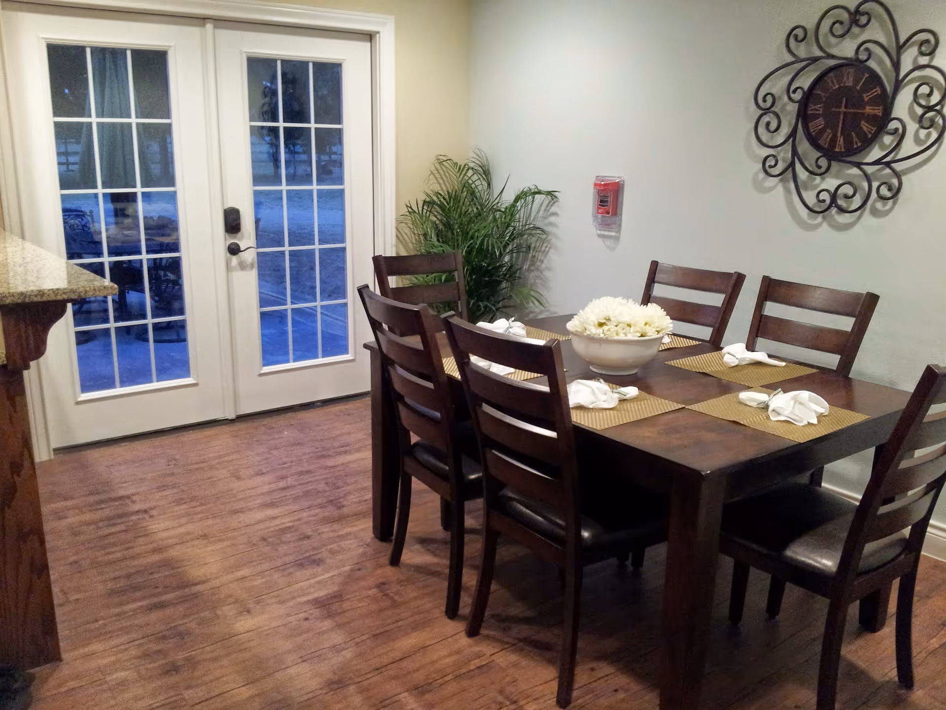 A dining area with a dark wooden table set for six with placemats and folded napkins. A decorative bowl with white flowers is in the center of the table. Behind the table is a wall with a large ornate clock and a fire alarm. To the left, there are double glass doors leading outside, and a green potted plant is placed in the corner.
