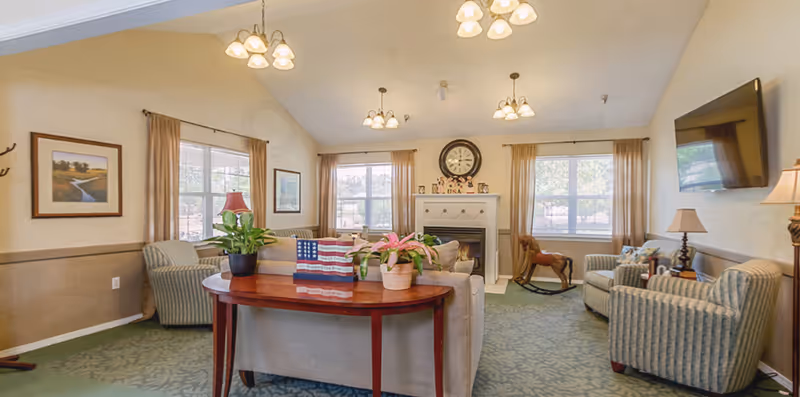 A cozy senior living facility common area with multiple armchairs and a sofa arranged around a fireplace. The room has large windows with beige curtains, a wall-mounted TV, a wooden rocking horse, and a table with potted plants. The walls are decorated with framed pictures and the carpet is green with a subtle pattern.