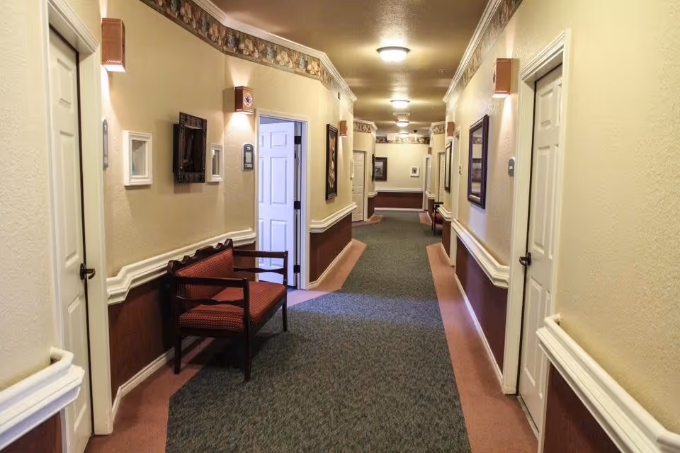 A well-lit indoor hallway in a senior living facility with beige walls, maroon lower wall paneling, and green carpet runner. Several closed white doors line both sides of the hallway, with framed pictures and wall sconces providing decoration. A wooden bench with red upholstery is placed along the left wall. The hallway extends into the distance with ceiling lights evenly spaced along the ceiling.