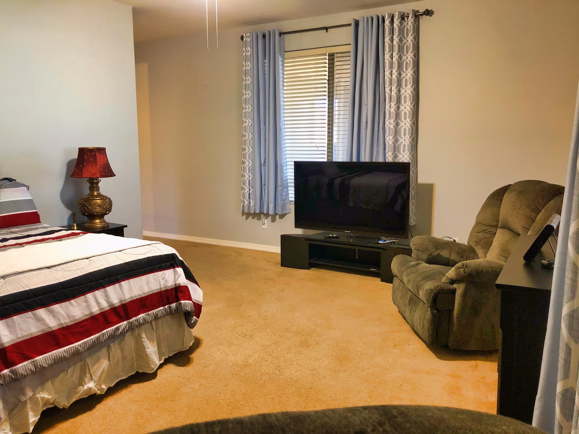 A cozy bedroom with beige carpet, a bed with a striped red, white, and black comforter, a decorative lamp on a nightstand, a large flat-screen TV on a black TV stand, a green recliner chair, and a window with blue and patterned curtains.
