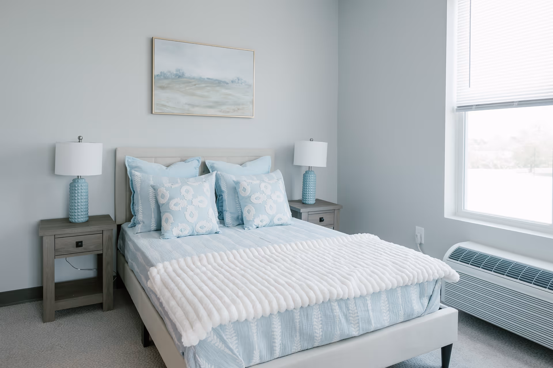 A neatly made bed with light blue and white bedding and multiple pillows in a bedroom. There are two matching bedside tables with blue lamps on each side of the bed. A framed landscape painting hangs on the wall above the bed. A large window with blinds is on the right side, and a heating/cooling unit is below the window.