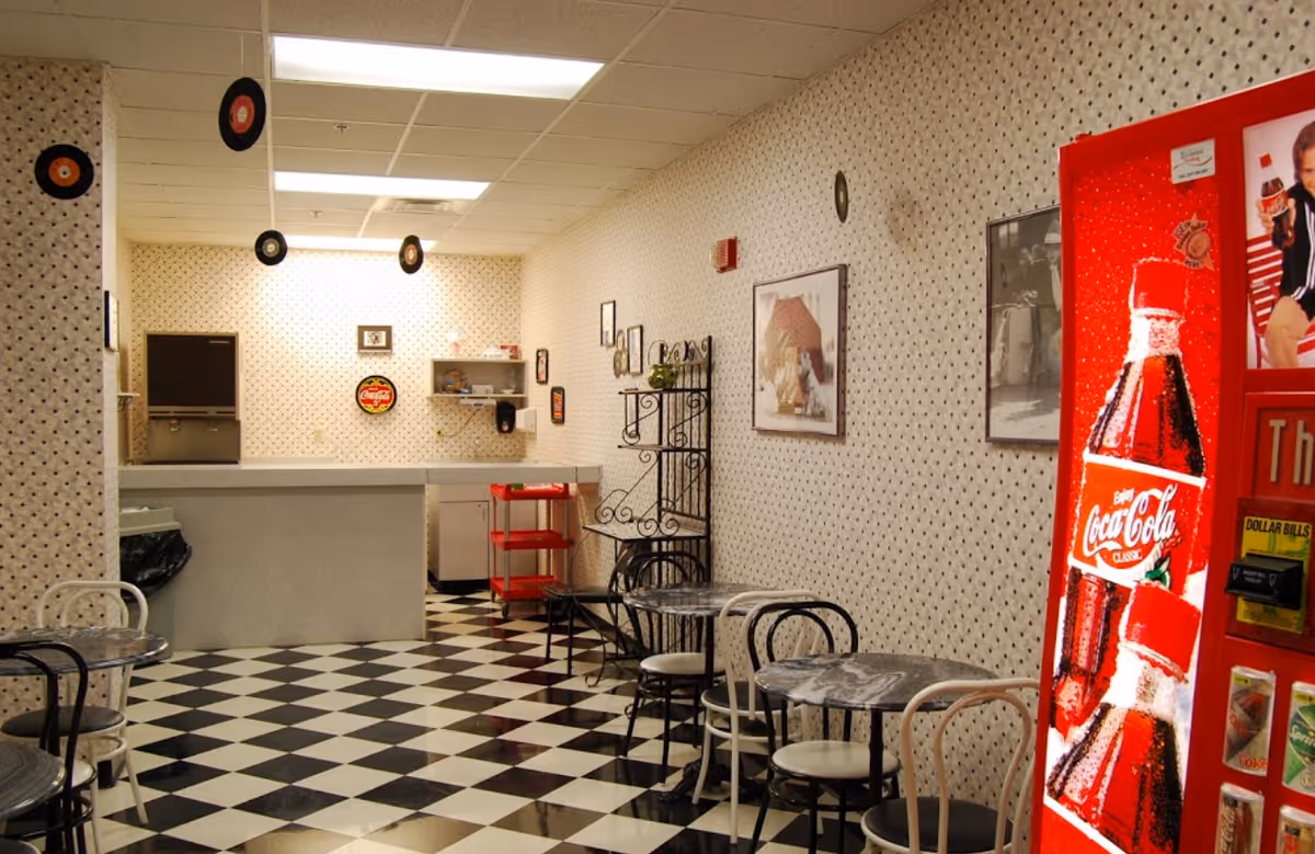 A retro-style dining area with black and white checkered floor tiles, small round tables with black and white chairs, patterned wallpaper, and a red Coca-Cola vending machine on the right. The back wall has a counter with a soda fountain machine, a red cart, and various framed pictures and decorations, including hanging vinyl records from the ceiling.
