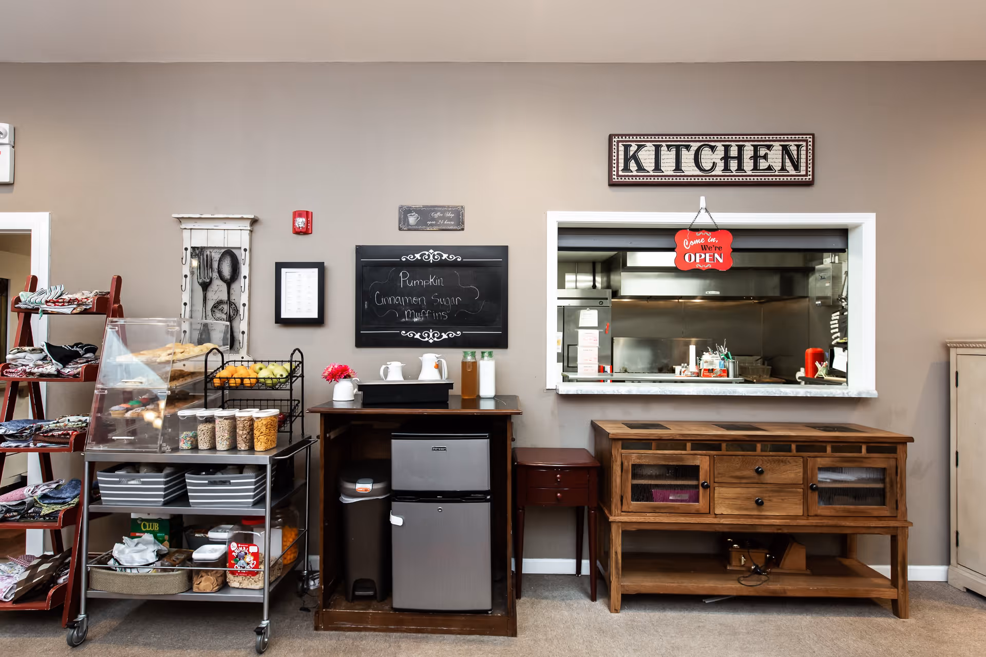 Interior view of a kitchen serving area in a senior living facility with a window opening to the kitchen. The wall above the window has a sign that says 'KITCHEN' and a red hanging sign that says 'Come in, We're OPEN'. Below the window is a wooden cabinet. To the left, there is a small refrigerator, a trash bin, a table with pitchers and bottles, a chalkboard with the menu listing Pumpkin Cinnamon Sugar Muffins, and a metal cart with snacks and baked goods. A red ladder-style shelf with folded cloths is also visible on the far left.