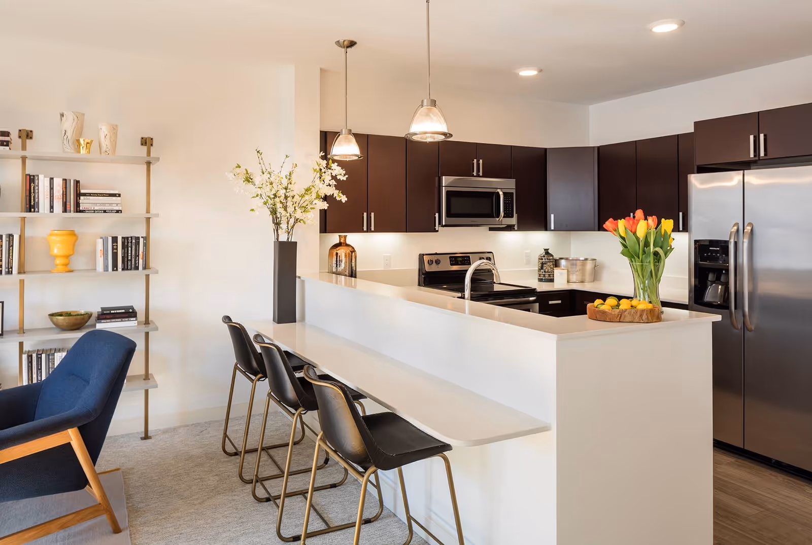 Modern kitchen with dark brown cabinets, stainless steel appliances including a refrigerator, stove, and microwave. A white countertop island with a built-in sink and three black bar stools. A vase with orange and yellow tulips and a wooden bowl with lemons are on the counter. To the left, there is a blue armchair and a bookshelf with books and decorative items.
