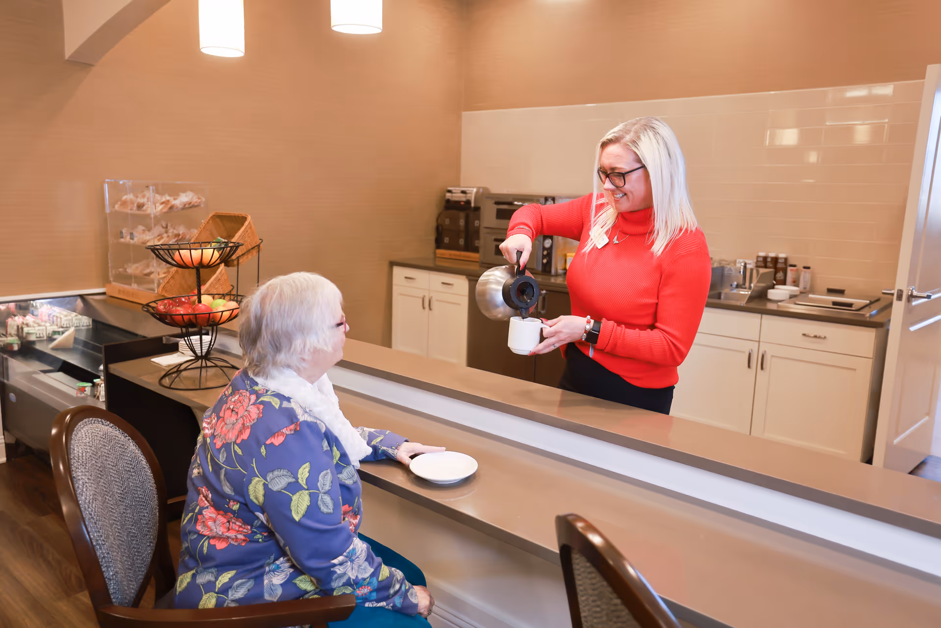 A woman in a red sweater pours coffee into a cup for an elderly woman seated at a counter in a kitchen-like area with cabinets and a display of fruit and pastries.