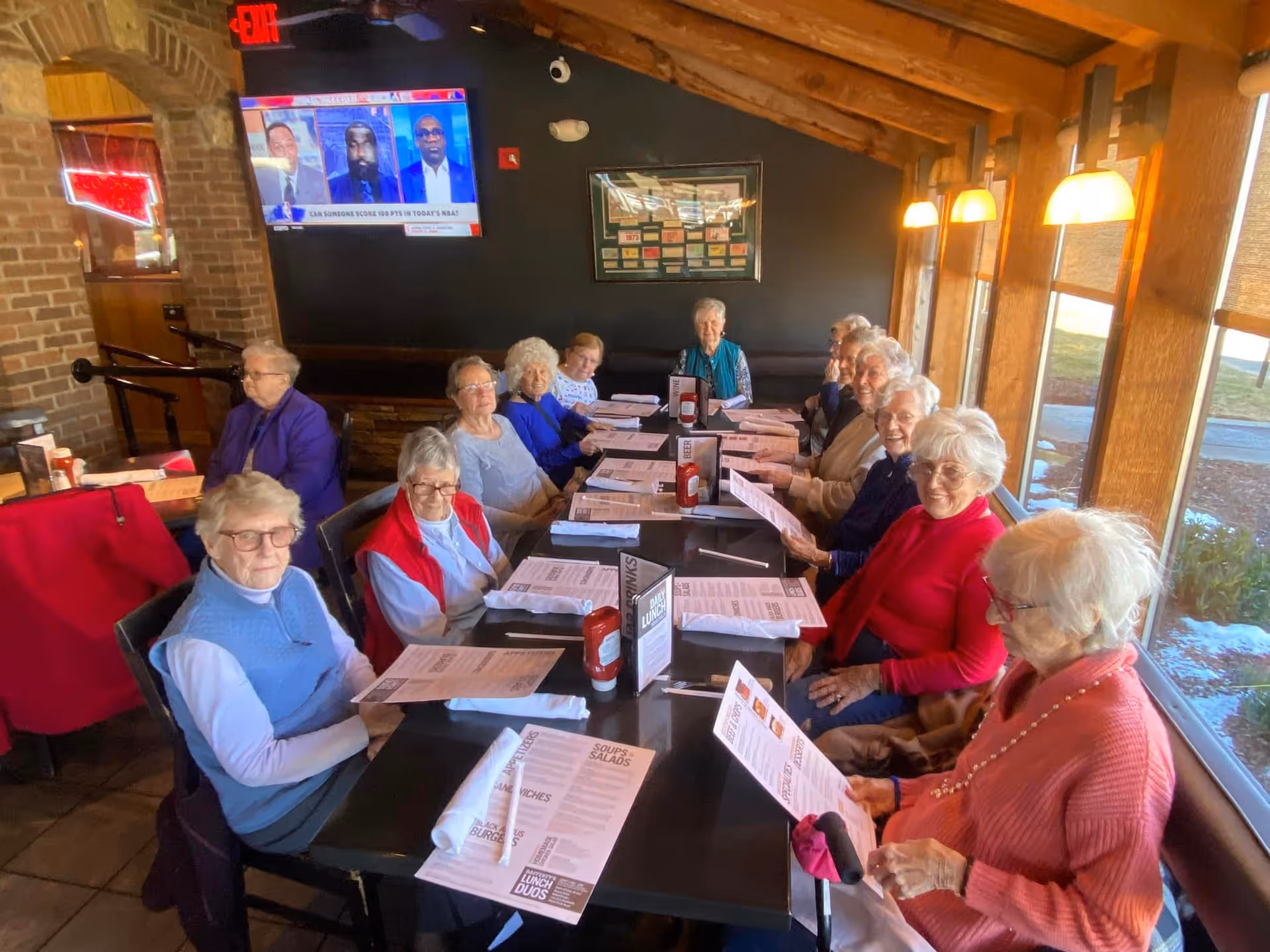 A group of elderly people sitting around a long dining table in a restaurant, each holding menus. The room has large windows on one side letting in natural light, a brick archway, and a television mounted on the wall showing a news program.