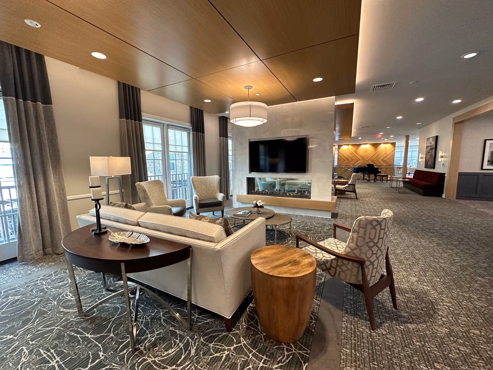 A spacious and modern living room area in a senior living facility featuring a beige sofa, patterned armchairs, a wooden side table, and a round coffee table. The room has large windows with curtains, a mounted flat-screen TV above a sleek fireplace, and a wooden ceiling panel with recessed lighting. In the background, there is a piano and additional seating along a carpeted hallway.