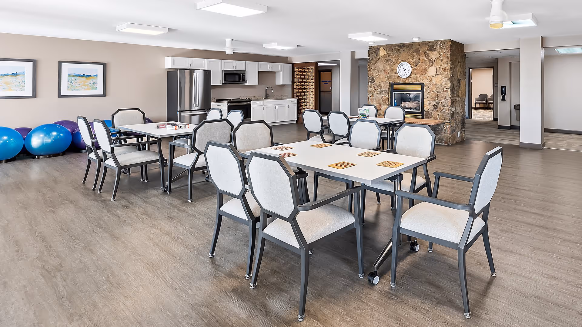 A spacious activity room with several tables and chairs arranged for games, a kitchenette with stainless steel refrigerator and microwave, exercise balls lined up against the wall, two framed landscape paintings, and a stone fireplace with a clock above it.