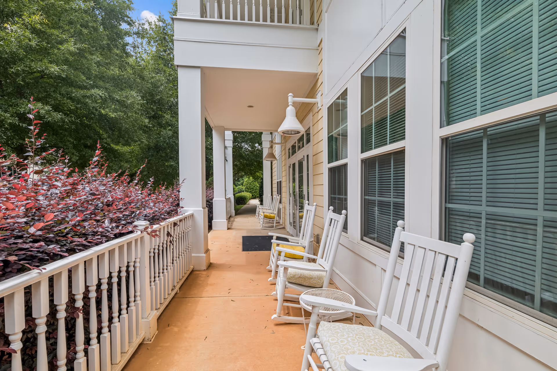 A covered outdoor porch area with white rocking chairs and small tables lined up along the wall of a building. The porch overlooks a garden with green trees and purple-leaved bushes. The building has large windows with closed blinds and white exterior walls.