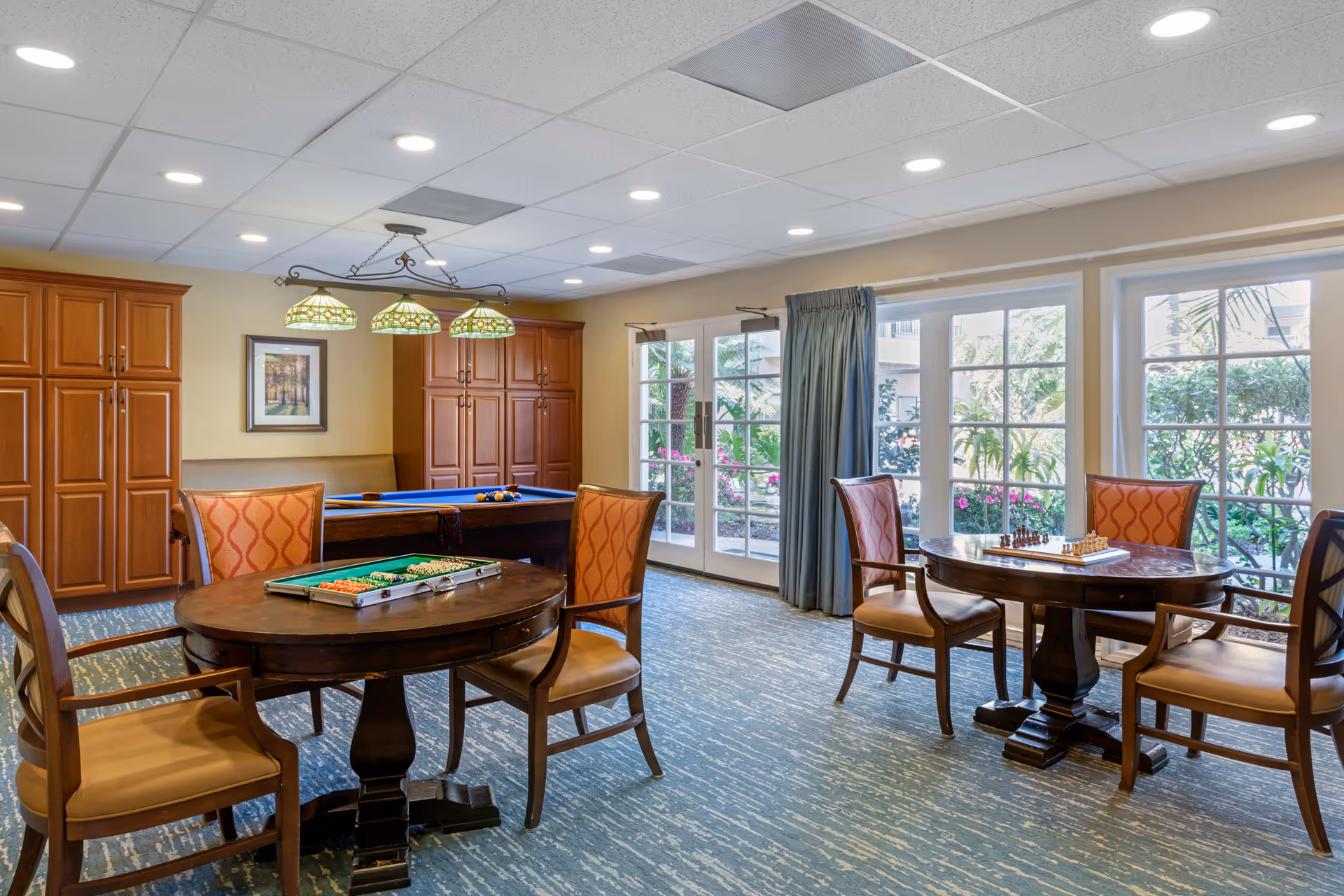 A bright and spacious game room with two round wooden tables, each surrounded by four chairs with orange patterned upholstery. One table has a backgammon set, and the other has a chess set. In the background, there is a pool table with a set of billiard balls and cues. Large windows and glass doors let in natural light and offer a view of greenery outside. Wooden cabinets line the back wall, and a decorative hanging light fixture is above the pool table.