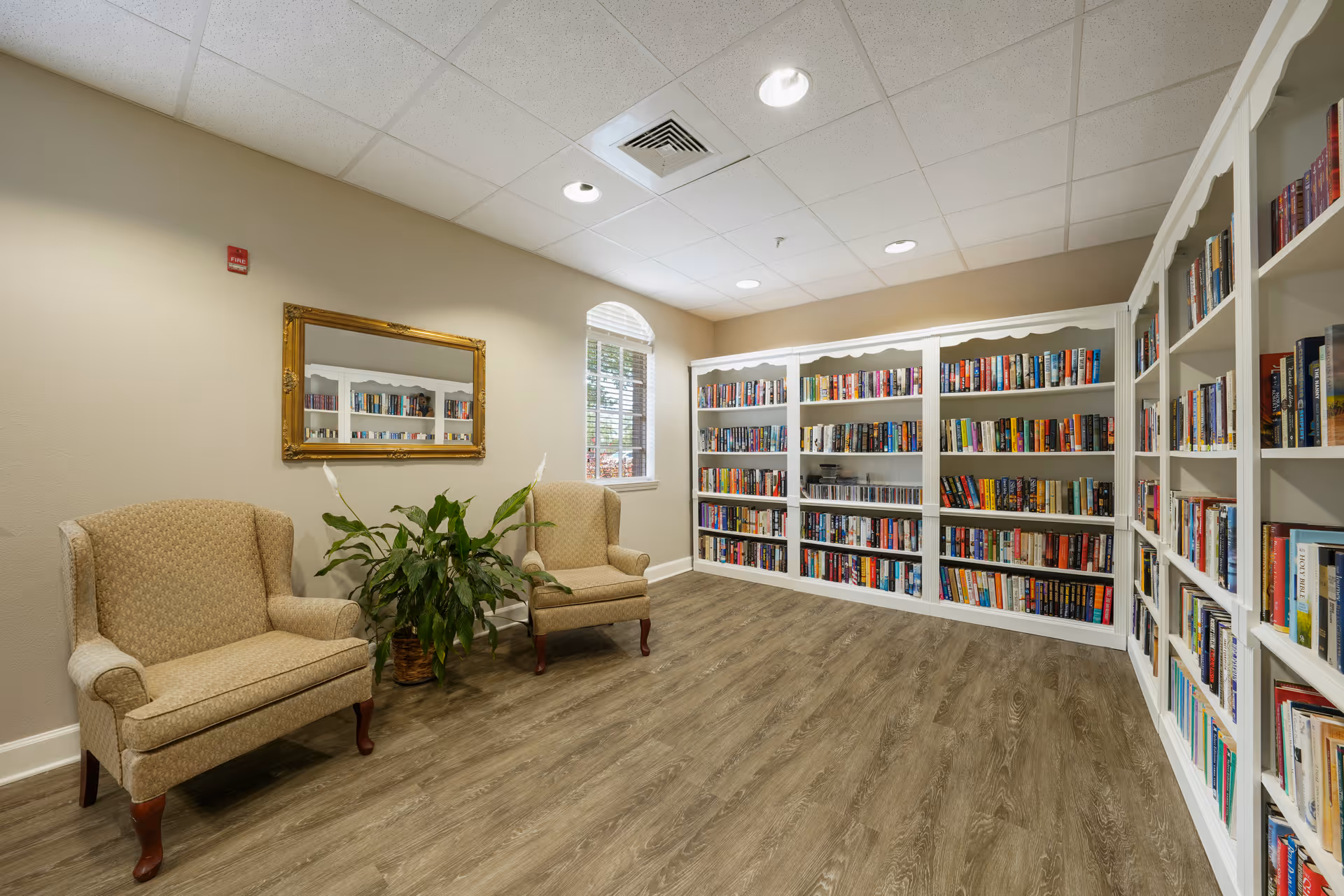 A cozy library room with two beige upholstered armchairs, a potted plant between them, a large gold-framed mirror on the wall, and white bookshelves filled with books along two walls. The room has wood flooring, a window with white blinds, and a white ceiling with recessed lighting.