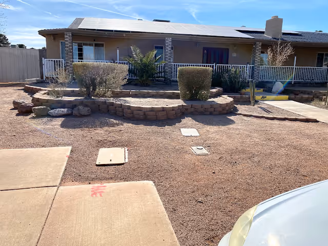 Exterior view of a single-story building with a covered porch, solar panels on the roof, and a landscaped front yard with bushes and gravel. A concrete walkway and driveway are visible in the foreground under a clear blue sky.