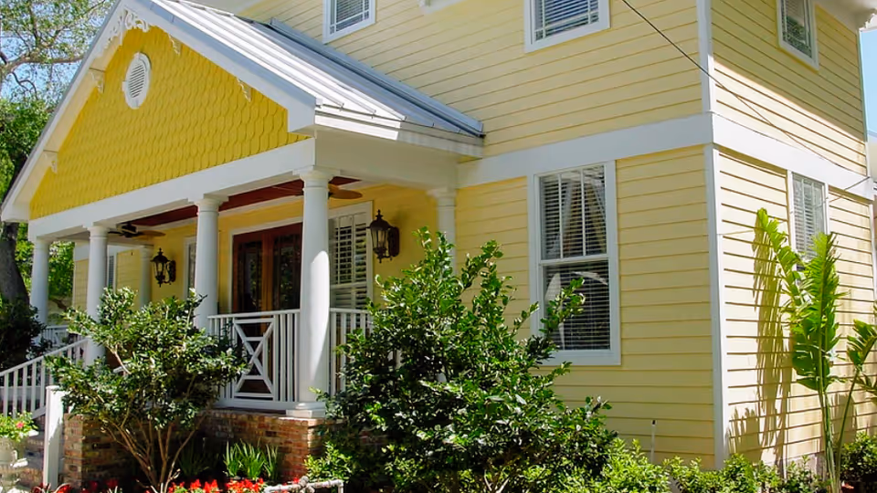 Exterior view of a yellow two-story house with white trim and columns supporting a covered porch. The porch has a white railing and two wall-mounted lanterns. There are green bushes and plants in front of the house, and the sky is clear and blue.