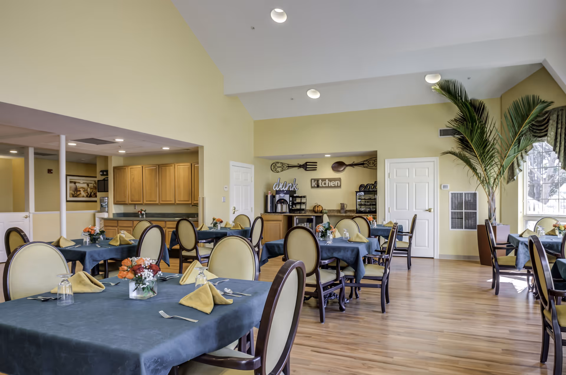 A bright dining room with several tables covered in dark blue tablecloths, each set with yellow napkins, silverware, and small floral centerpieces. The room has light yellow walls, a wooden floor, and a high ceiling with recessed lighting. There is a kitchen area in the background with wooden cabinets and a counter, decorated with large fork and spoon wall art and signs that say 'drink', 'kitchen', and 'eat'. A large potted palm plant is near a window with green curtains.