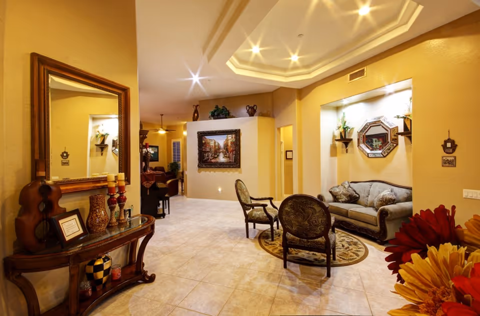A warmly lit interior common area with beige walls and tiled floor. The space features a decorative wooden console table with candles, vases, and a violin-shaped ornament. A large mirror hangs above the table. In the background, there is a sitting area with a sofa and two upholstered chairs arranged around a small round rug. The walls are adorned with framed artwork and decorative shelves with plants. The ceiling has recessed lighting and a tray ceiling design.