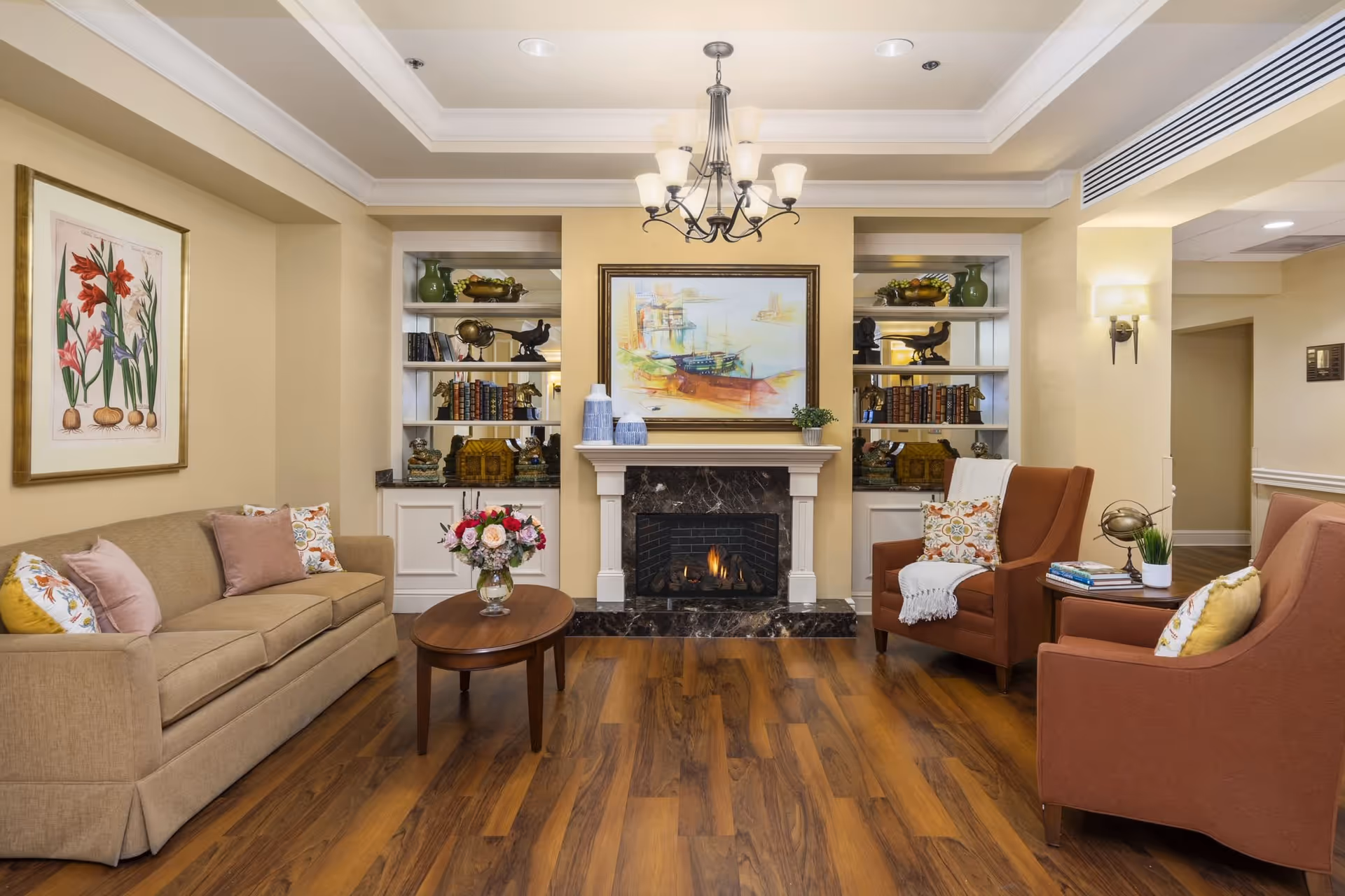 A cozy living room with a beige sofa and two rust-colored armchairs arranged around a wooden coffee table with a vase of flowers. The room features a fireplace with a marble surround, built-in white shelves filled with books and decorative items, a chandelier hanging from the ceiling, and a framed painting above the fireplace. The walls are painted a warm beige, and the floor is wood.