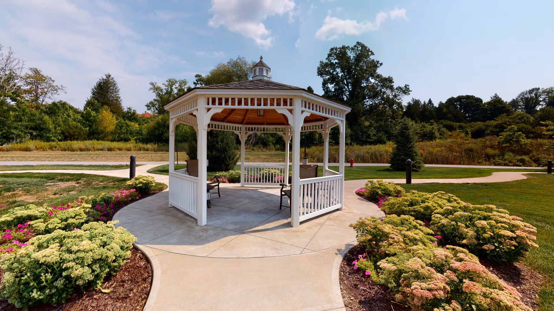 A white wooden gazebo with a shingled roof situated on a concrete circular platform surrounded by landscaped flower beds with green and pink plants. The gazebo is located in a garden area with paved walking paths and trees in the background under a partly cloudy sky.