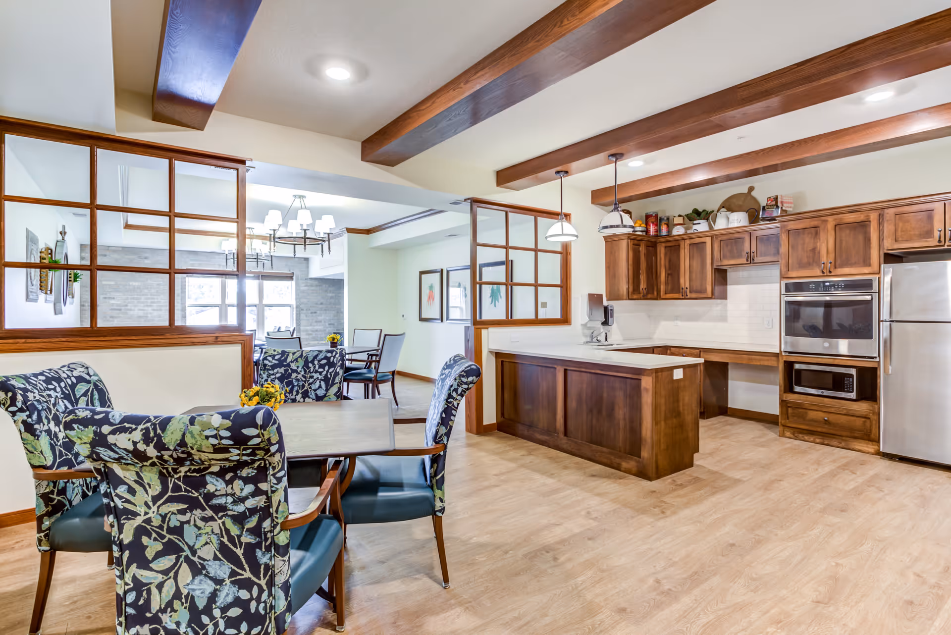 A bright and spacious senior living facility kitchen and dining area with wooden cabinetry, stainless steel appliances, and a small dining table with floral upholstered chairs. The kitchen features a countertop island and pendant lighting, with a dining room visible through wooden-framed glass partitions.
