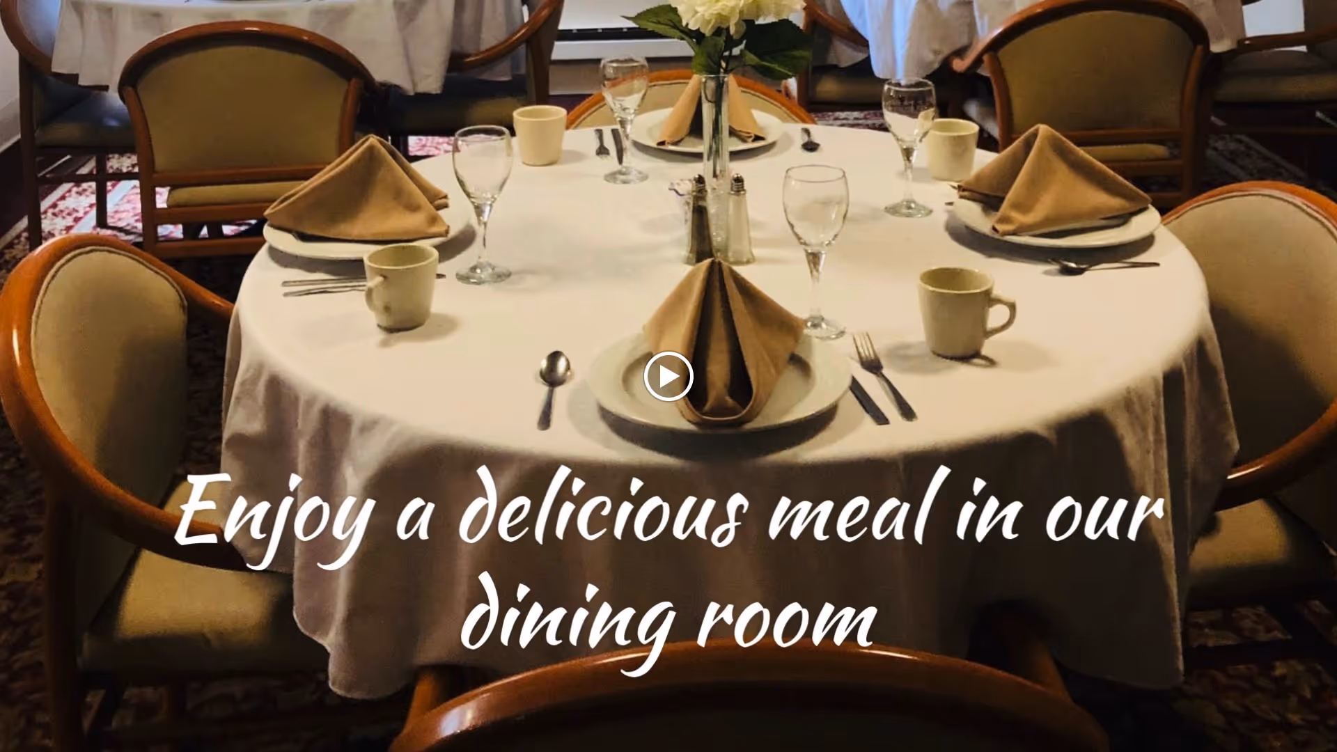 A round dining table set with a white tablecloth, beige folded napkins on plates, cups, glasses, and silverware in a dining room with upholstered chairs and a floral centerpiece.