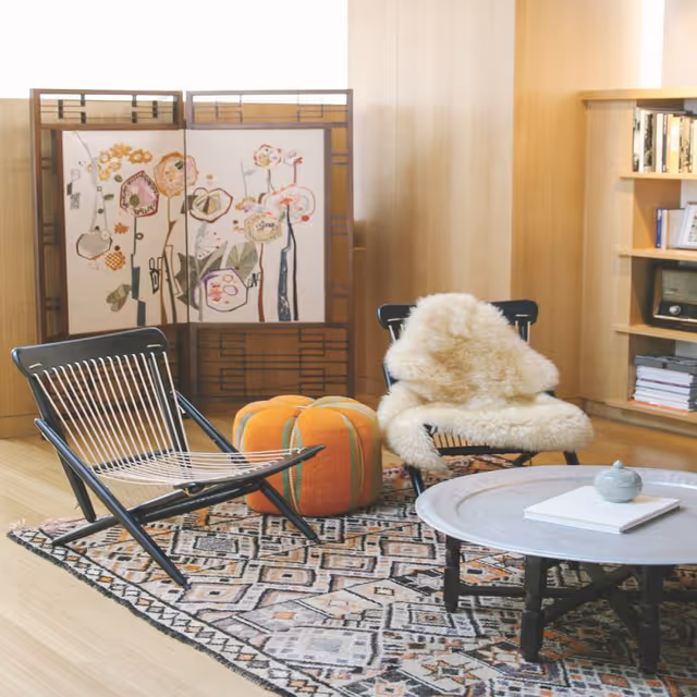 A cozy living room area with two modern black chairs, one draped with a fluffy white throw, an orange and green striped pouf, a round gray coffee table with a small decorative item and a book, a patterned rug, a wooden room divider with floral artwork, and a built-in wooden bookshelf filled with books and decor.