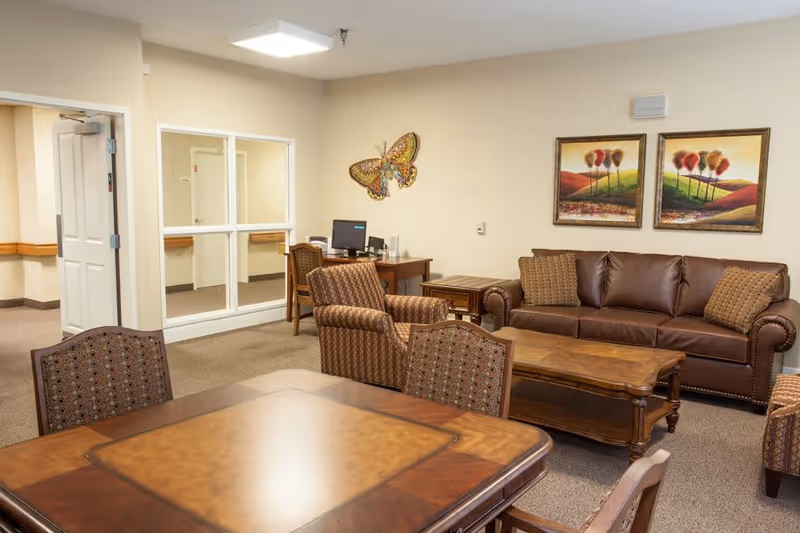 A cozy senior living facility common area with a brown leather sofa, two patterned armchairs, a wooden coffee table, and a wooden dining table with chairs. The walls are decorated with two landscape paintings and a large butterfly wall art. A desk with a computer is visible in the background near a window and an open door.