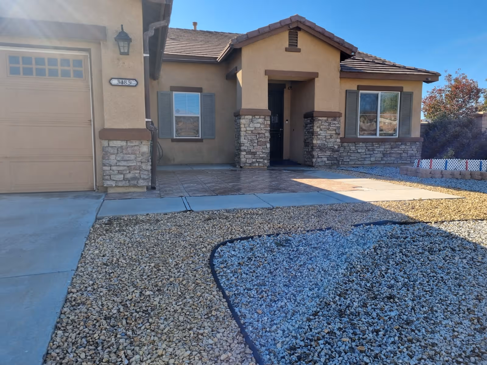 Front exterior view of a single-story house with a beige stucco finish, stone accents around the entrance and windows, a tiled porch, and a gravel yard with a small garden area on the right side.