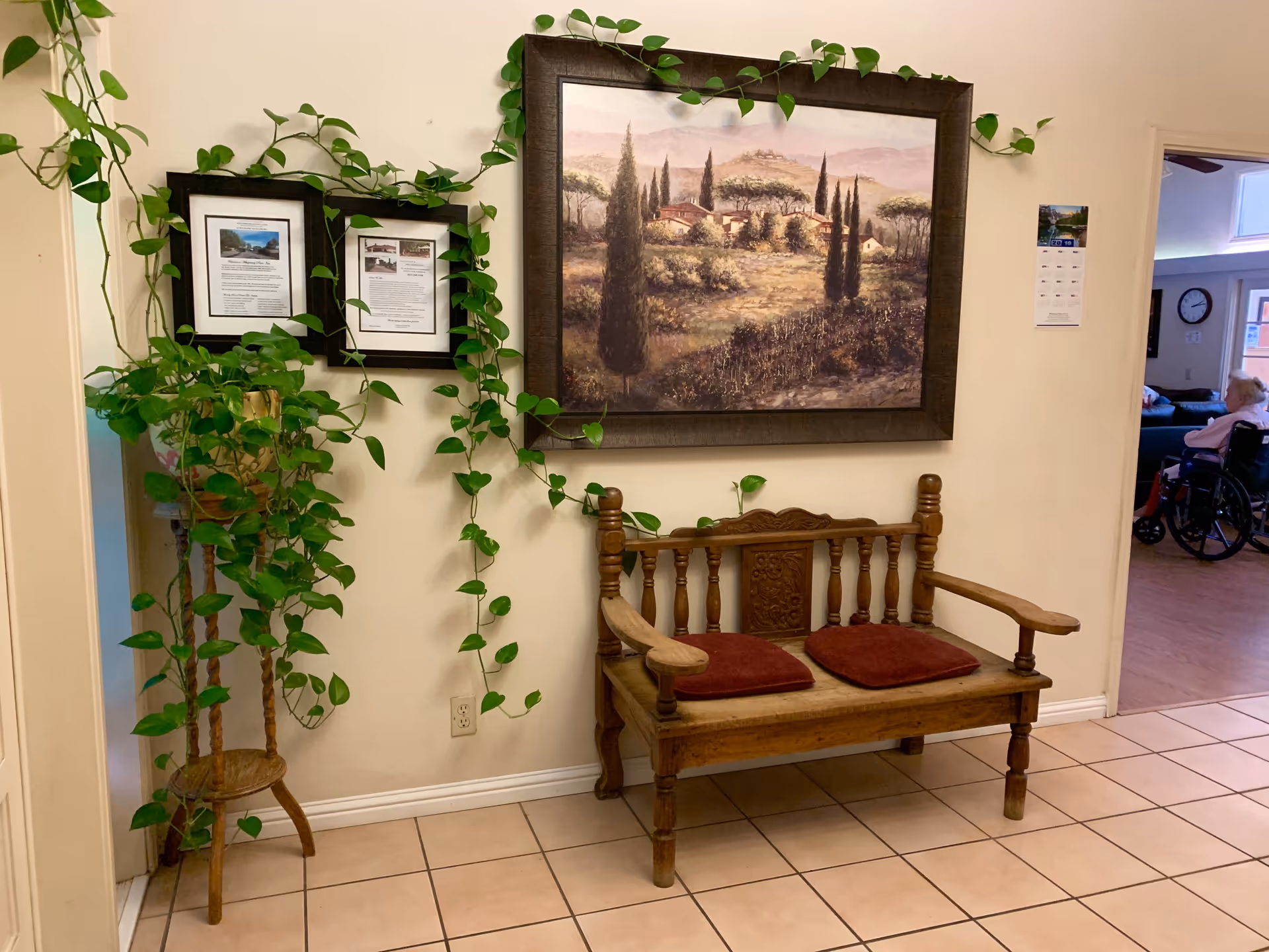 Wooden bench with red cushions beneath a large framed landscape painting and trailing potted plant in a tiled interior hallway of a senior living facility.
