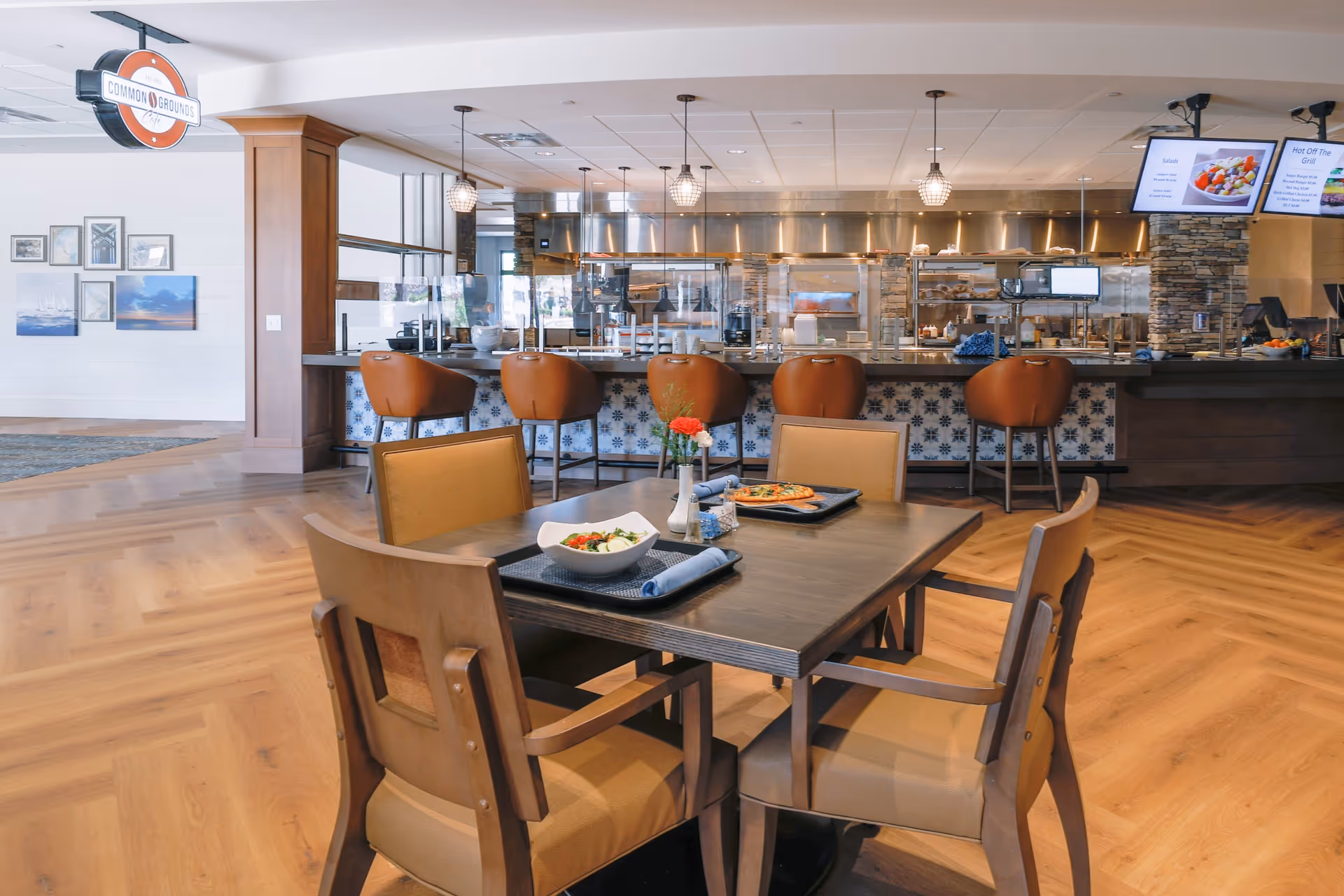 Interior view of a modern dining area at Town Center at Covenant Living of Cromwell featuring a wooden table with four chairs, two trays with food including a salad and a pizza, a vase with flowers, and a counter with five brown bar stools in front of an open kitchen. The kitchen has stainless steel appliances and hanging pendant lights. A sign above the counter reads 'Common Grounds Cafe'.