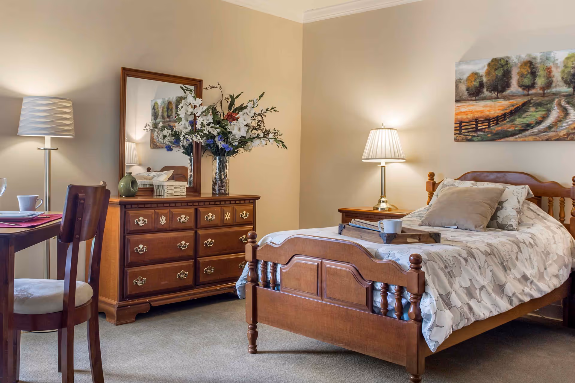A cozy bedroom with a wooden bed featuring a floral-patterned comforter and pillows. A wooden dresser with a large mirror and a vase of flowers sits against the wall. A table with a chair and a cup is visible on the left side. Two lamps provide warm lighting, and a landscape painting hangs above the bed.