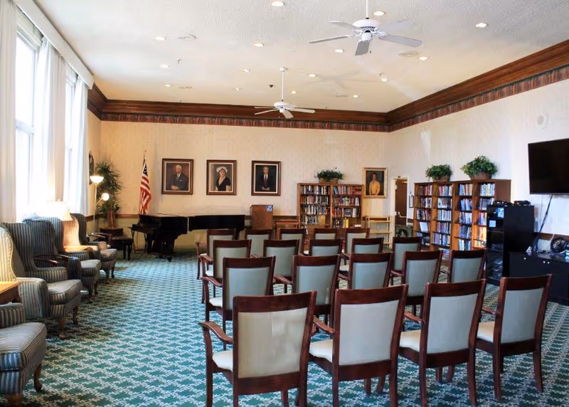 Carpeted community room with rows of chairs facing a piano, bookshelves, and a TV.