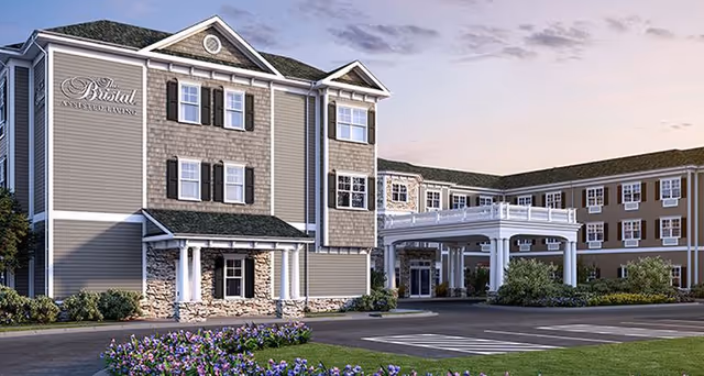 Exterior view of The Bristal Assisted Living at West Babylon building during dusk, featuring a multi-story structure with gray siding, white trim, and stone accents. The entrance has a covered driveway with white columns, surrounded by landscaped greenery and purple flowers.