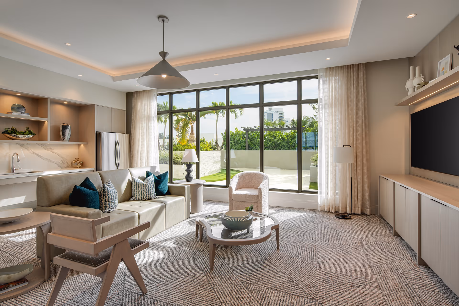 A bright and modern living room with large floor-to-ceiling windows showing an outdoor patio with palm trees. The room features a beige sofa with decorative pillows, a wooden armchair, a round glass coffee table with decorative items, a side table with a lamp, and a large flat-screen TV mounted on the wall. The space also includes a small kitchen area with a refrigerator, sink, and open shelves with decorative pieces.