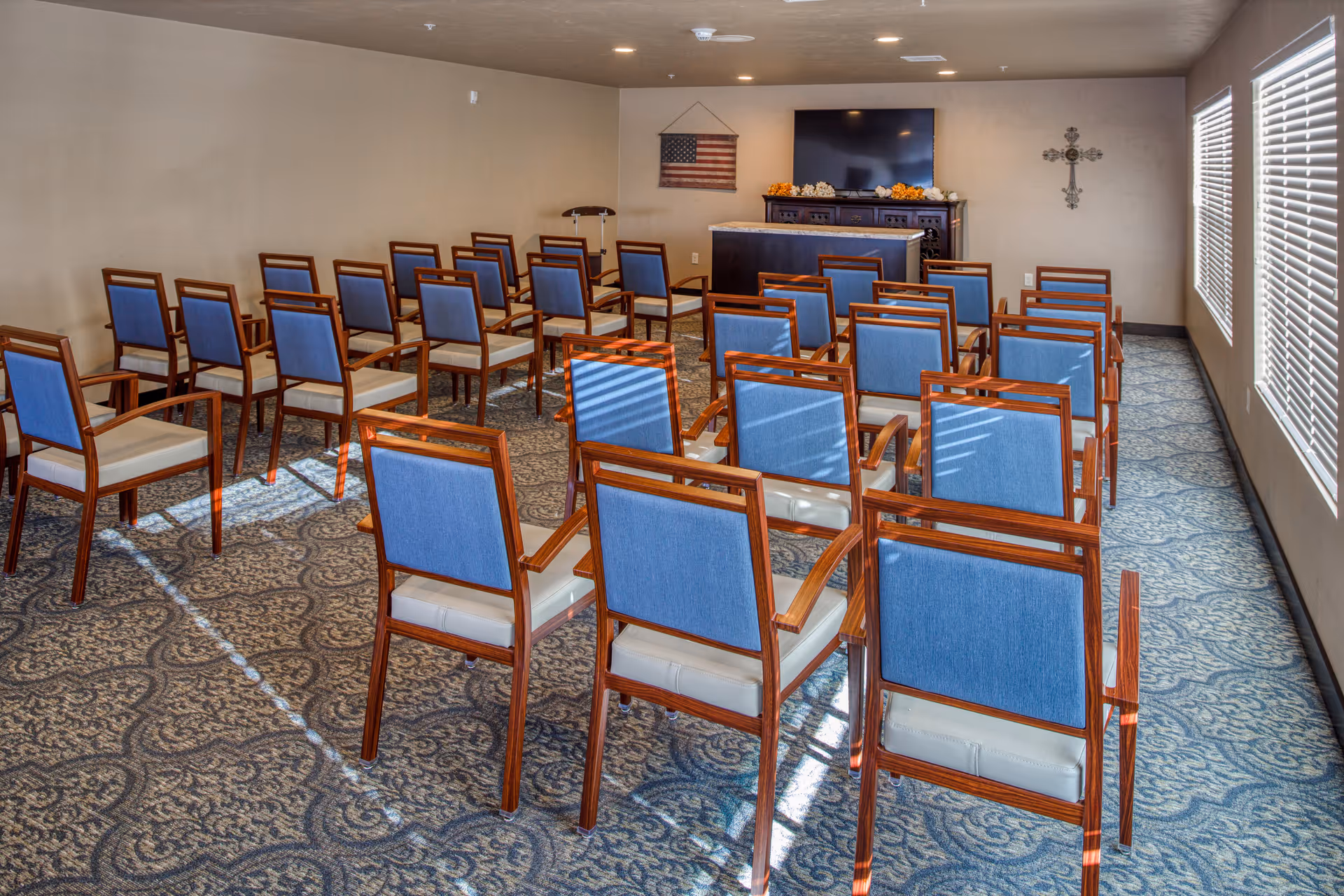 A small meeting or chapel room with rows of wooden chairs with blue backs and white seats arranged facing a podium and a TV mounted on the wall. An American flag hangs on the wall to the left of the TV, and a decorative cross is mounted on the wall to the right. The room has patterned carpet flooring and windows with blinds on the right side.