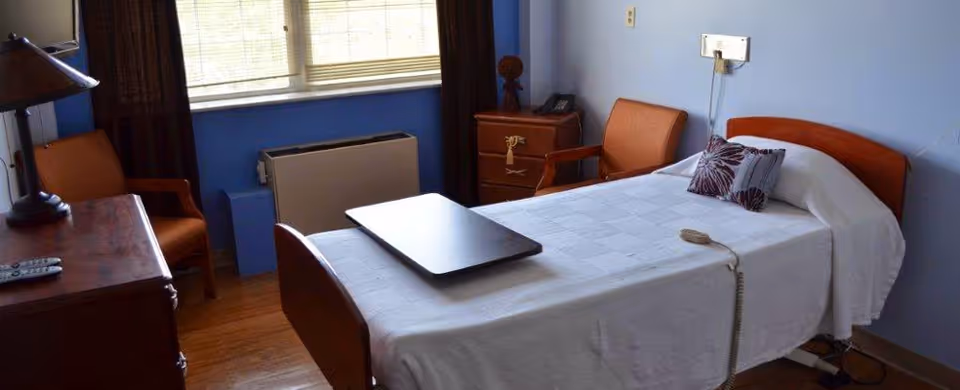 A simple nursing facility bedroom with a single bed, bedside table, chairs, and a window.