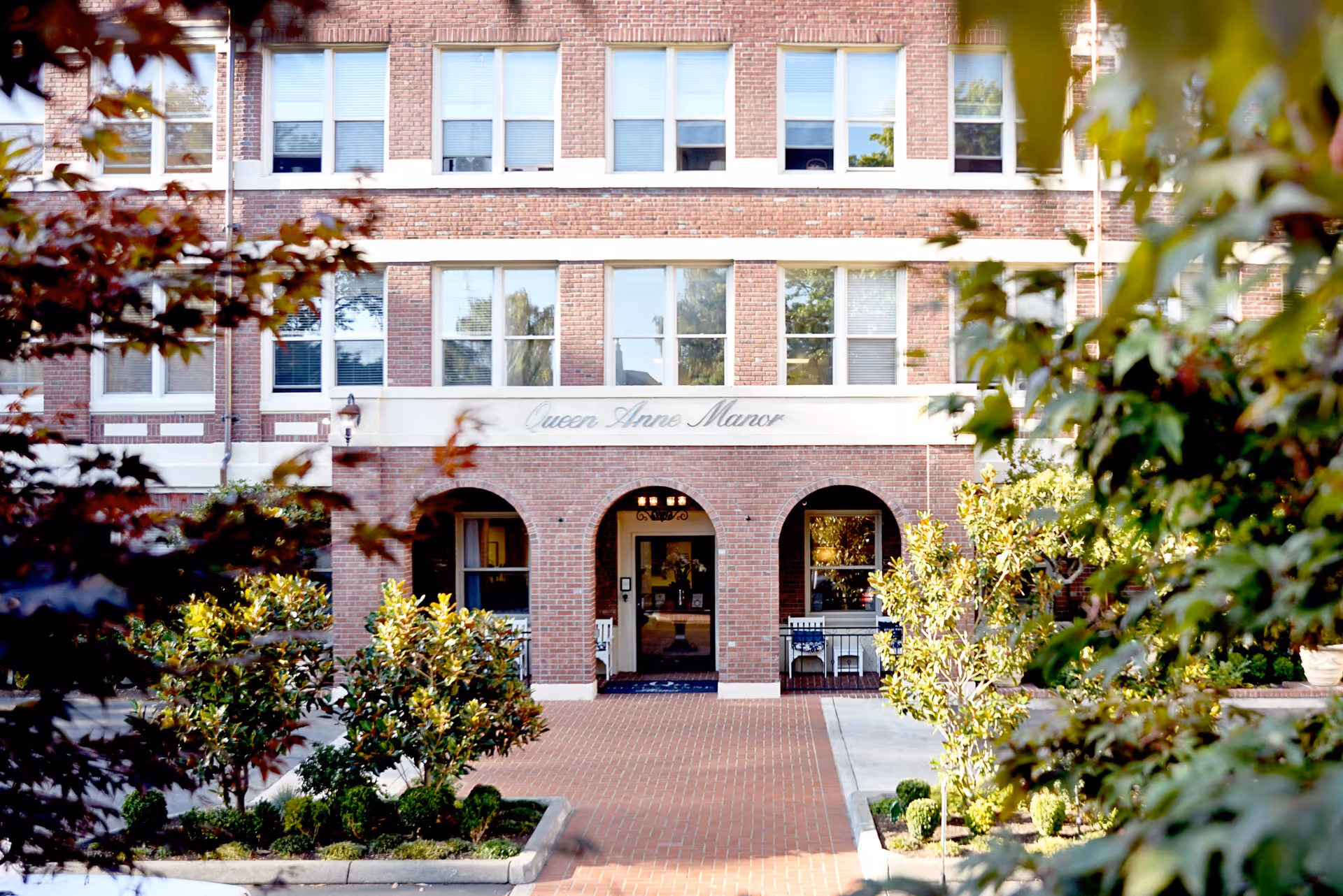 Front exterior view of a brick building named Queen Anne Manor, with multiple windows and an arched entrance. The entrance is framed by greenery and small trees along a brick walkway.