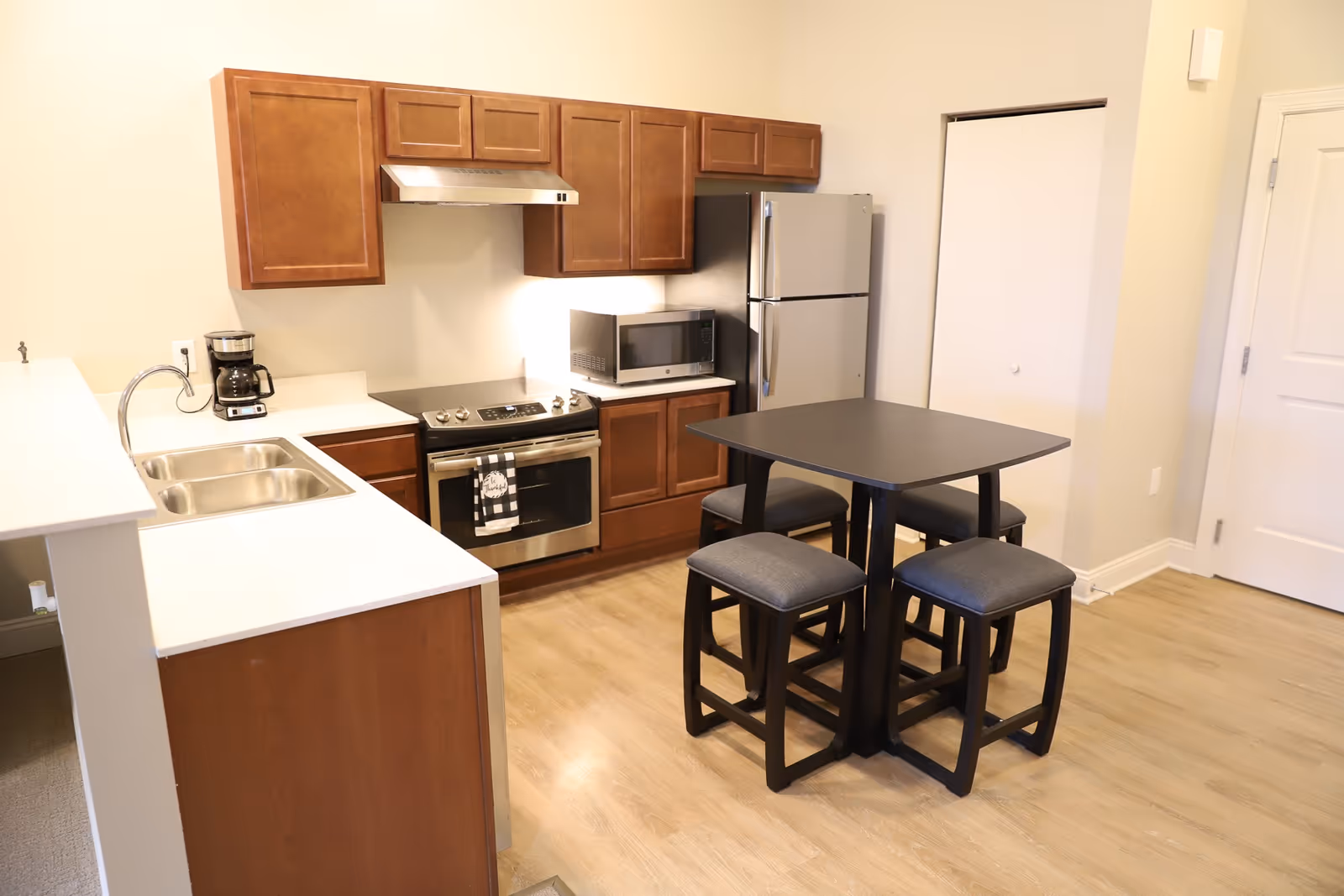 A modern kitchen with wooden cabinets, a stainless steel stove and refrigerator, a microwave, a coffee maker, a double sink, and a small dining table with four cushioned stools on a light wood floor.