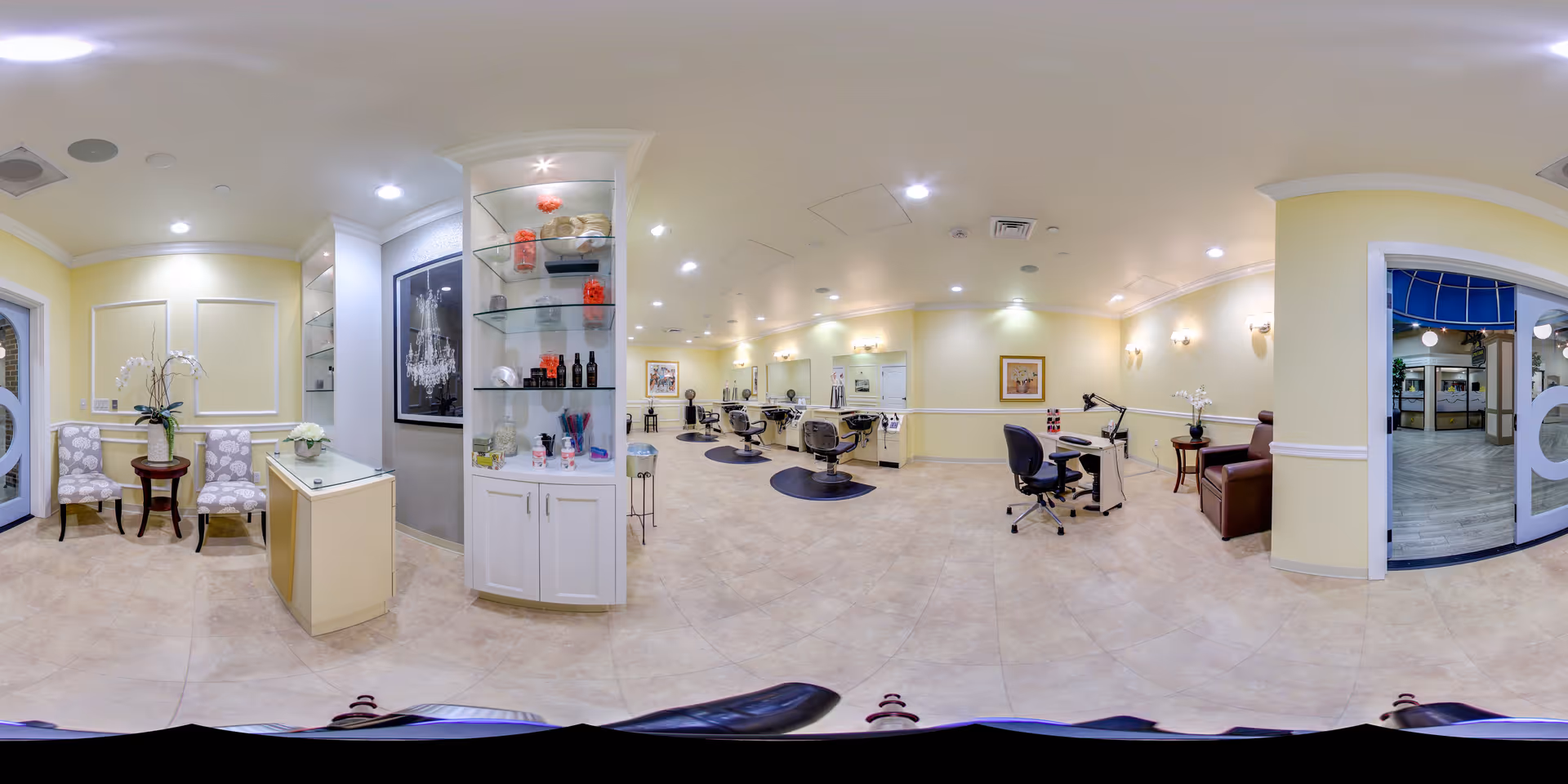 A spacious and well-lit salon area in a senior living facility with multiple styling chairs and mirrors along the walls. There is a small reception desk with decorative flowers, a glass display cabinet with hair care products, and a manicure station with a chair and lamp. The walls are painted light yellow with white trim, and the floor is tiled. Two upholstered chairs with a small table and a plant are near the entrance, and large glass doors lead to another area.