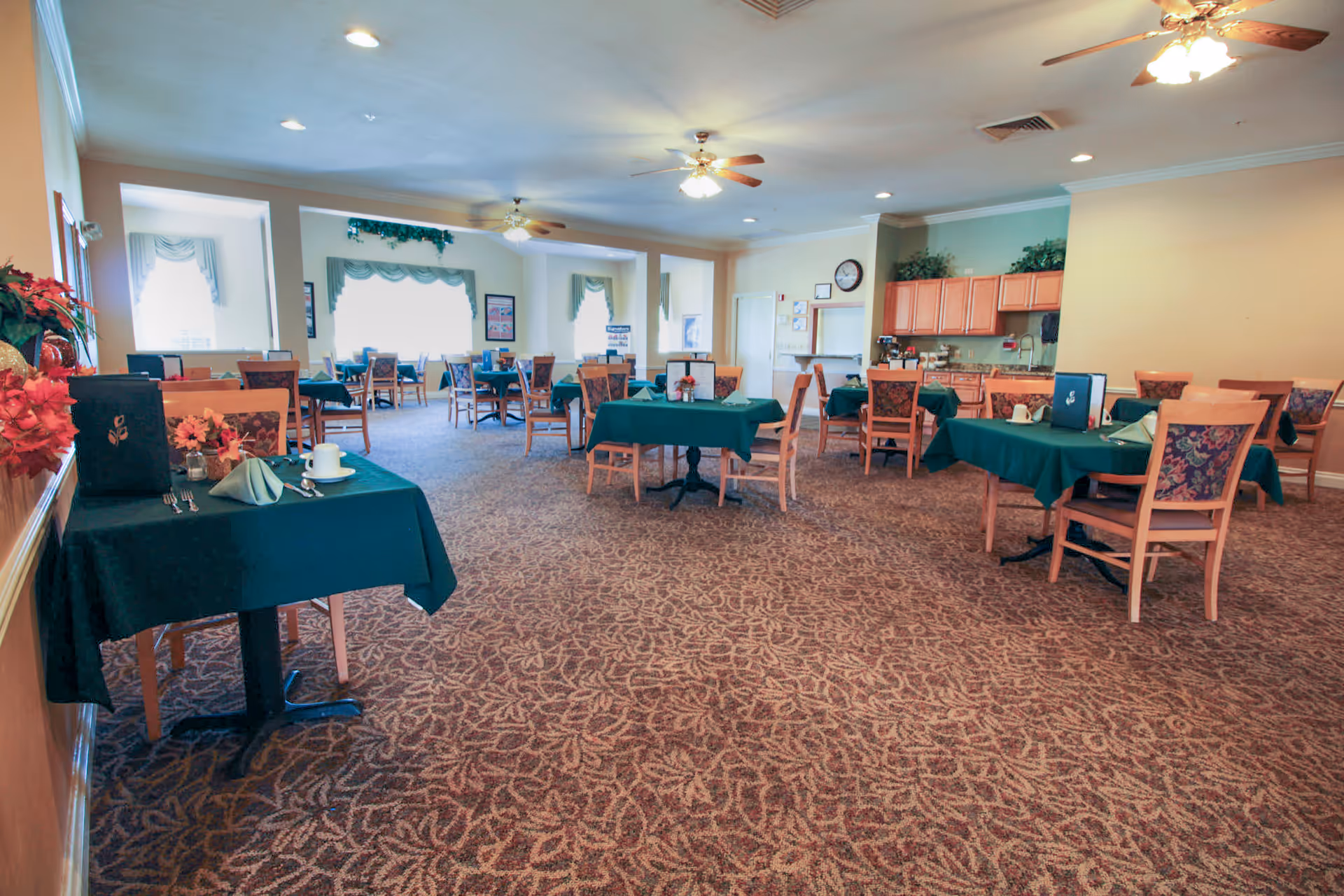 A spacious dining room with multiple tables covered in green tablecloths, each set with napkins, cups, and menus. The room has patterned carpet flooring, ceiling fans with lights, large windows with decorative curtains, and a small kitchen area with cabinets and a sink in the back.